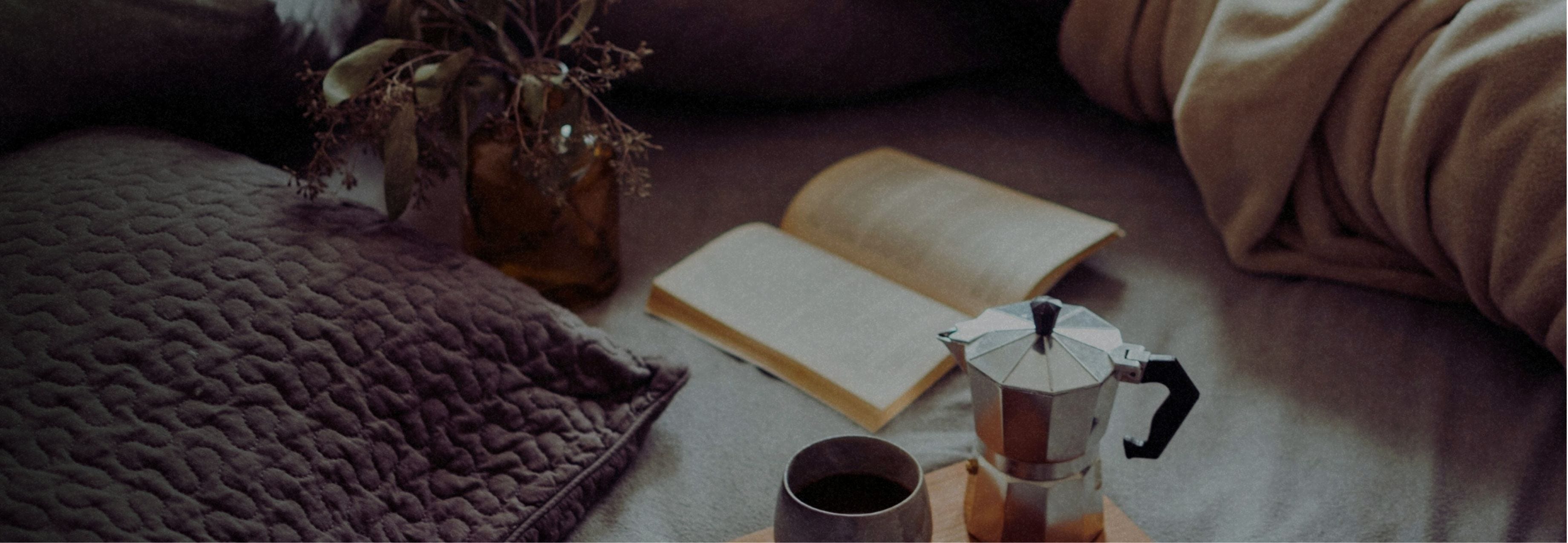 A softly lit bedside scene with an open notebook, a cup of tea, a textured throw blanket, and a small vase with dried stems, creating a calm, cozy atmosphere.