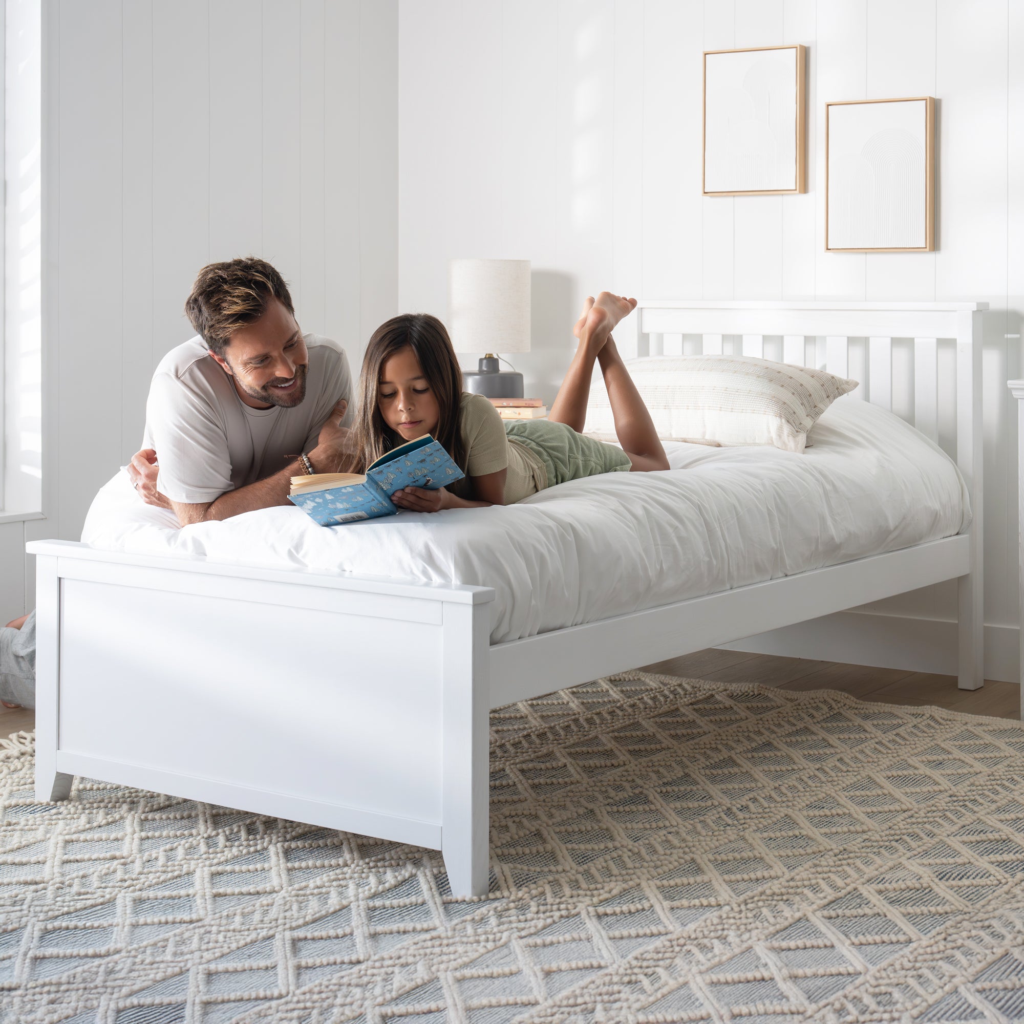 Man and child lying on a white bed reading a book in a bright bedroom. #size_Twin Platform #color_washed white