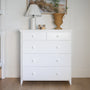 A white wooden five-drawer dresser with round white knobs, styled with a lamp, small decorative blocks, and a wicker vase  #color_washed white