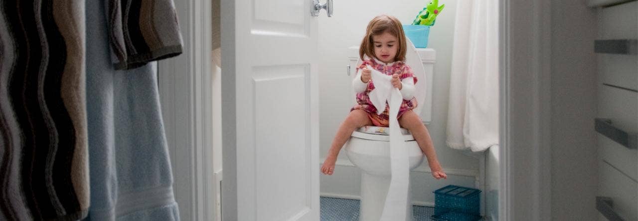 Toddler girl sitting on the toilet and unraveling a roll of toilet paper