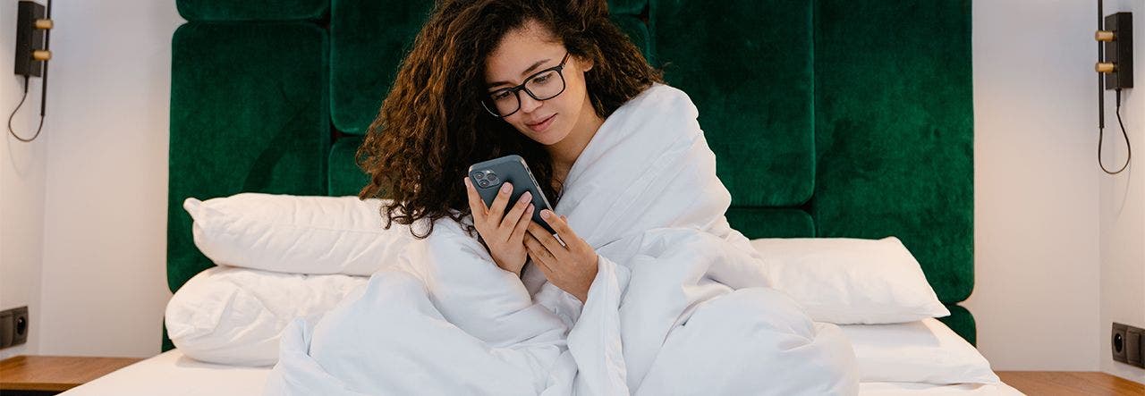 Woman sitting up in bed, wrapped in a blanket and looking at her phone