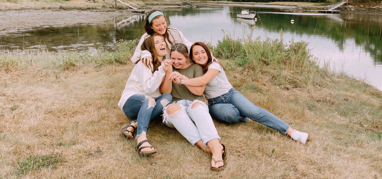 Four young women sitting beside a pond, hugging and smiling