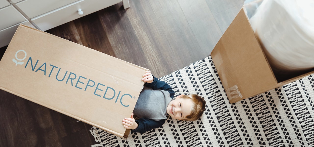 Smiling boy sliding our of a Naturepedic mattress box, roll-packed mattress nearby