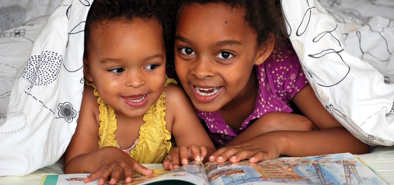 Two litte girls reading together under a blanket and giggling 