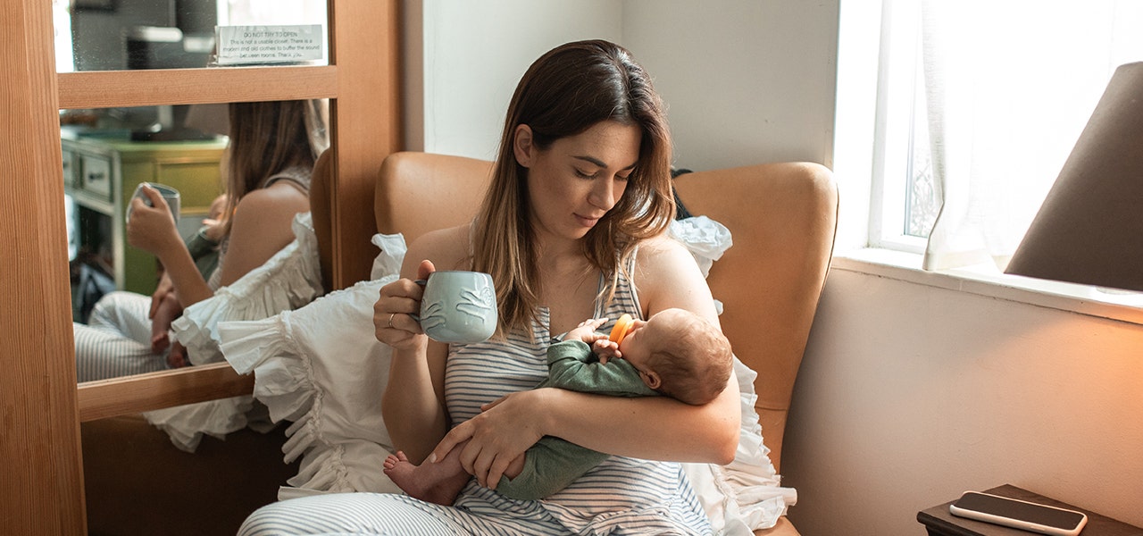 Mother sitting in nursery chair, holding her baby and drinking a warm beverage