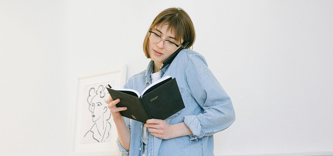 woman wearing glasses holding a book and talking on the phone